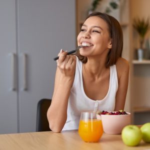 An image of a women eating and smiling. She has a bowl of food in front of her, orange juice and an apple.