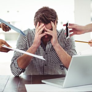 A man sitting by a laptop with his head in his hands, there are people pointing things like phones and papers from him in different directions
