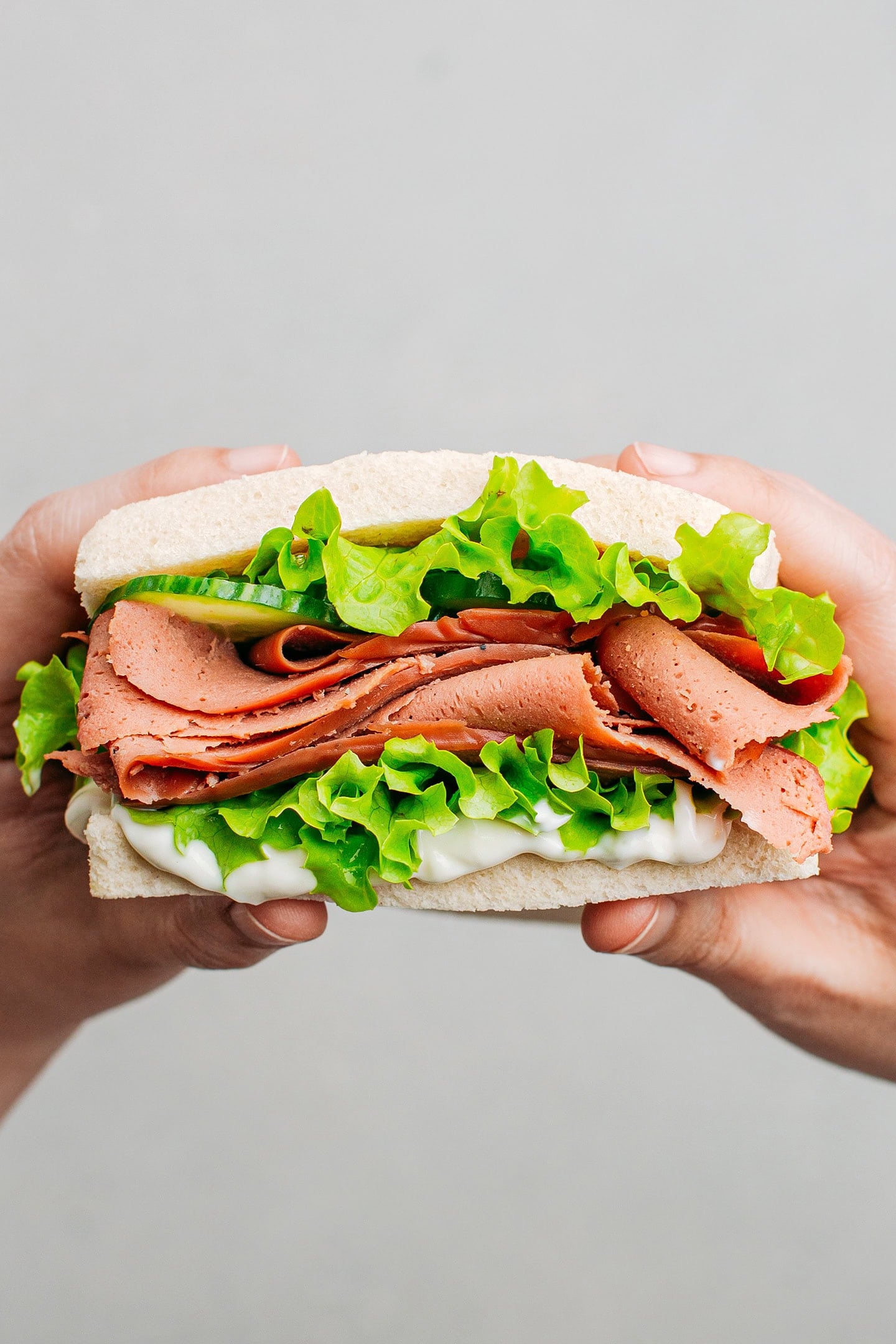 Holding a sandwich filled with vegan deli slices, mayo, cucumber, and lettuce.
